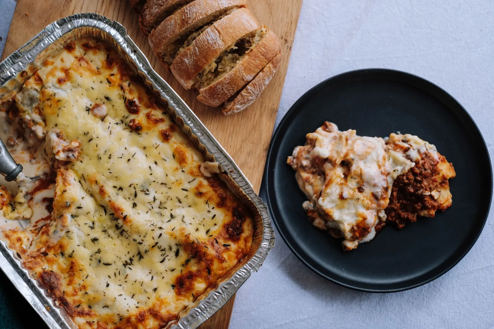 Delicious lasagna in foil container with plated portion and fresh bread